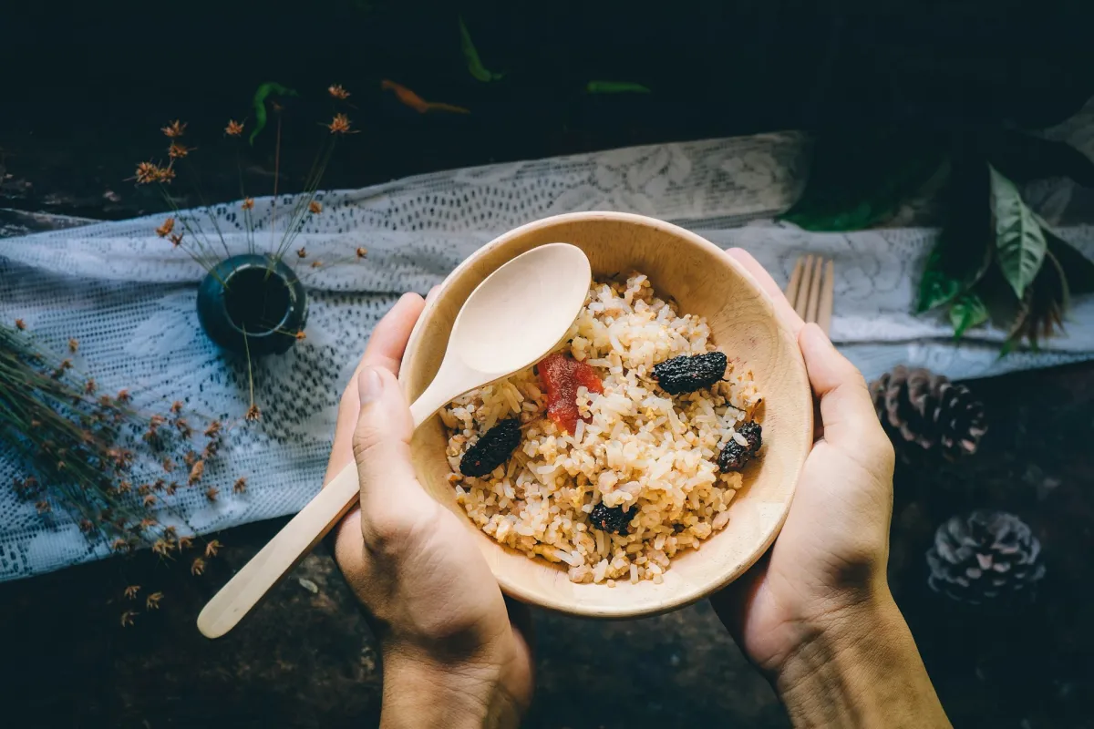 Manos sosteniendo un cuenco de madera con arroz, frutas secas y una cuchara de madera
