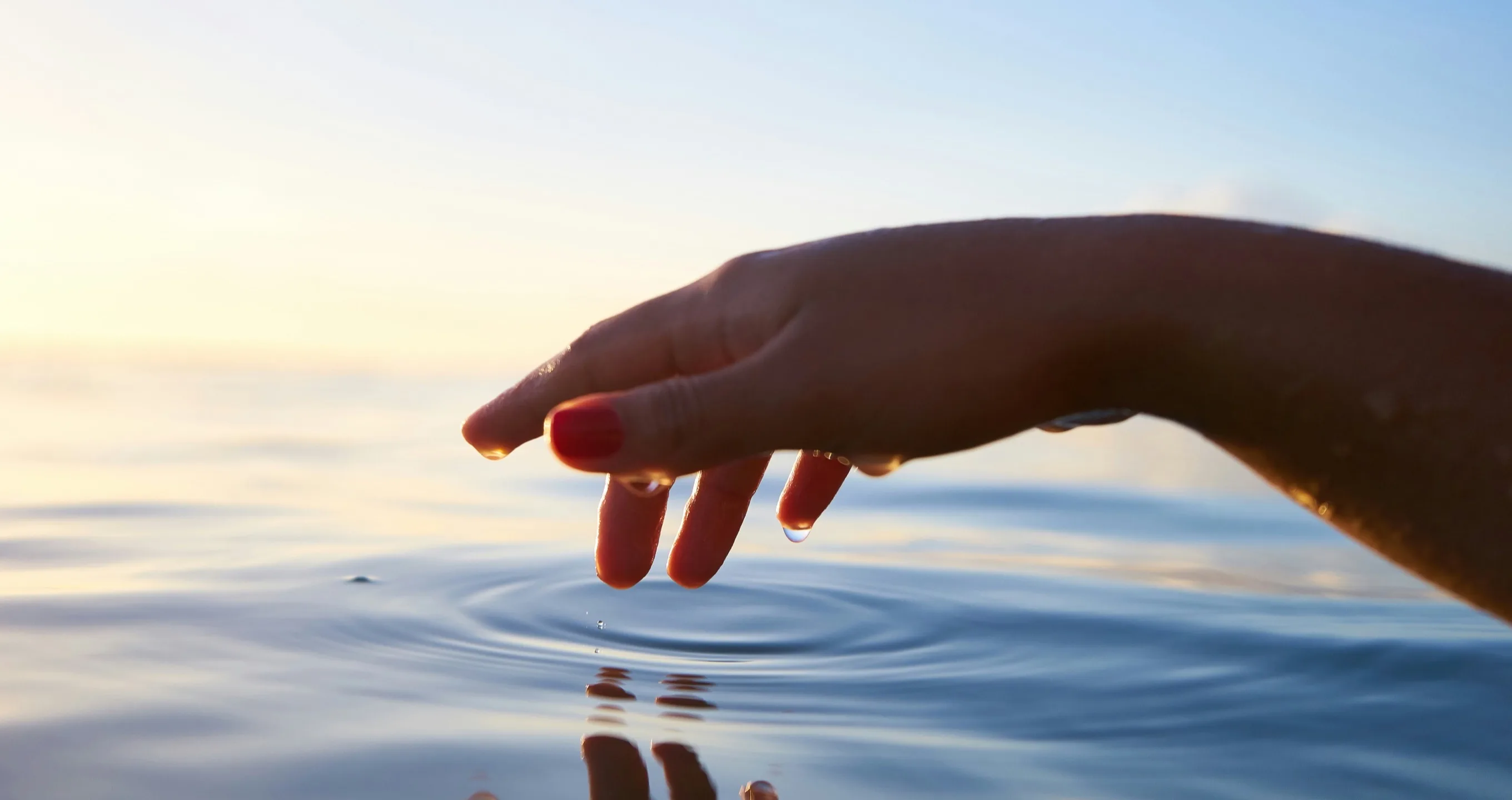 A hand reaching down to touch the calm surface of water at sunset, sending out ripples