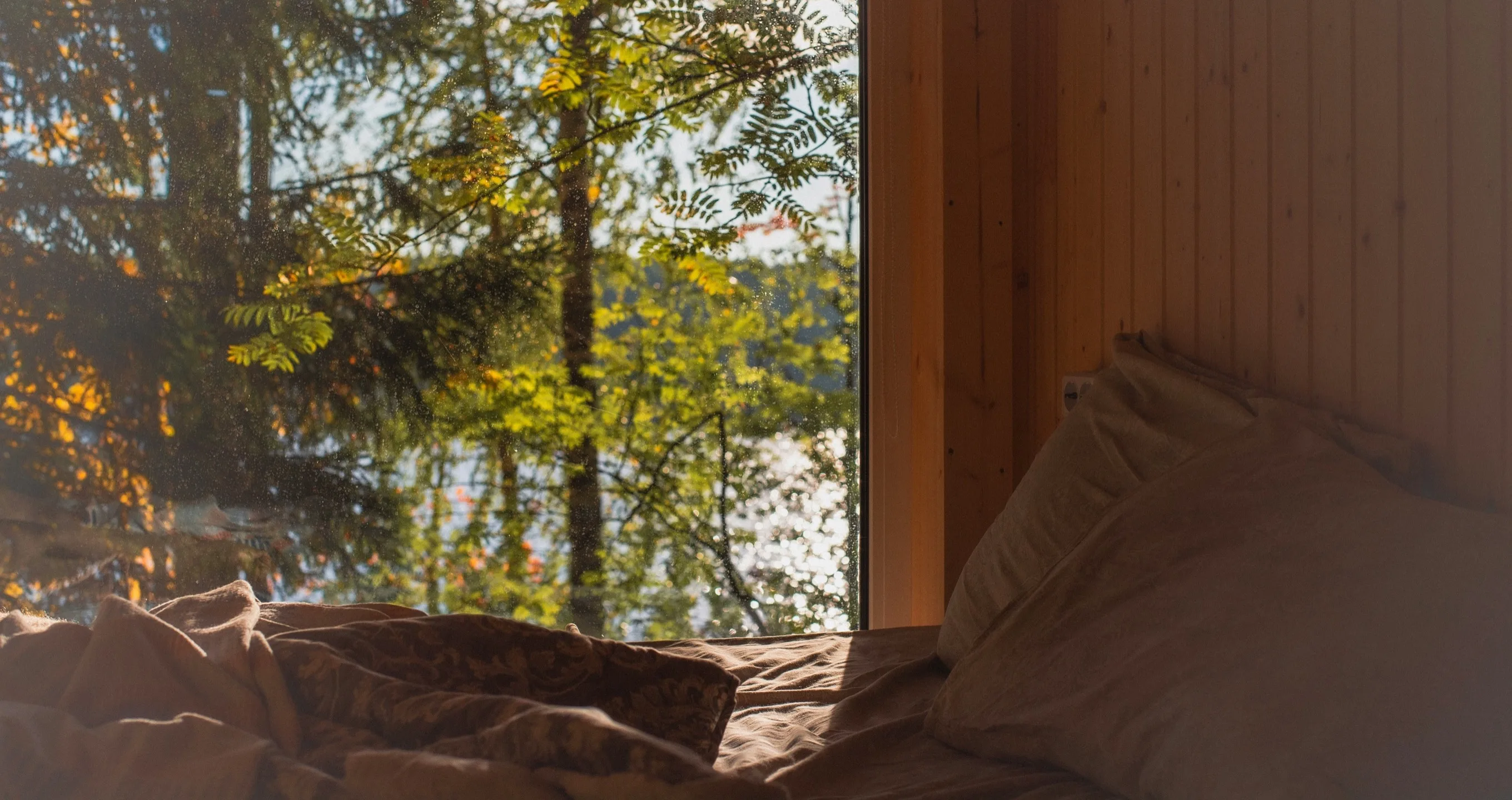 An unmade bed beside a large window looking out onto a sunlit autumn forest and lake — a quiet morning scene of rest and slow waking.